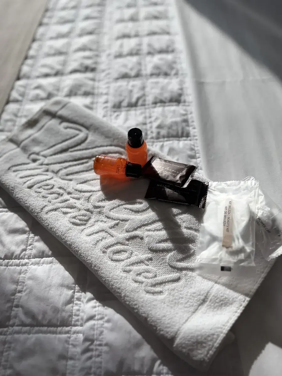 Toalha branca com "Violeta Meaípe Hotel" bordado, sobre a cama, com xampu, condicionador, sabonete em barra e kit de beleza