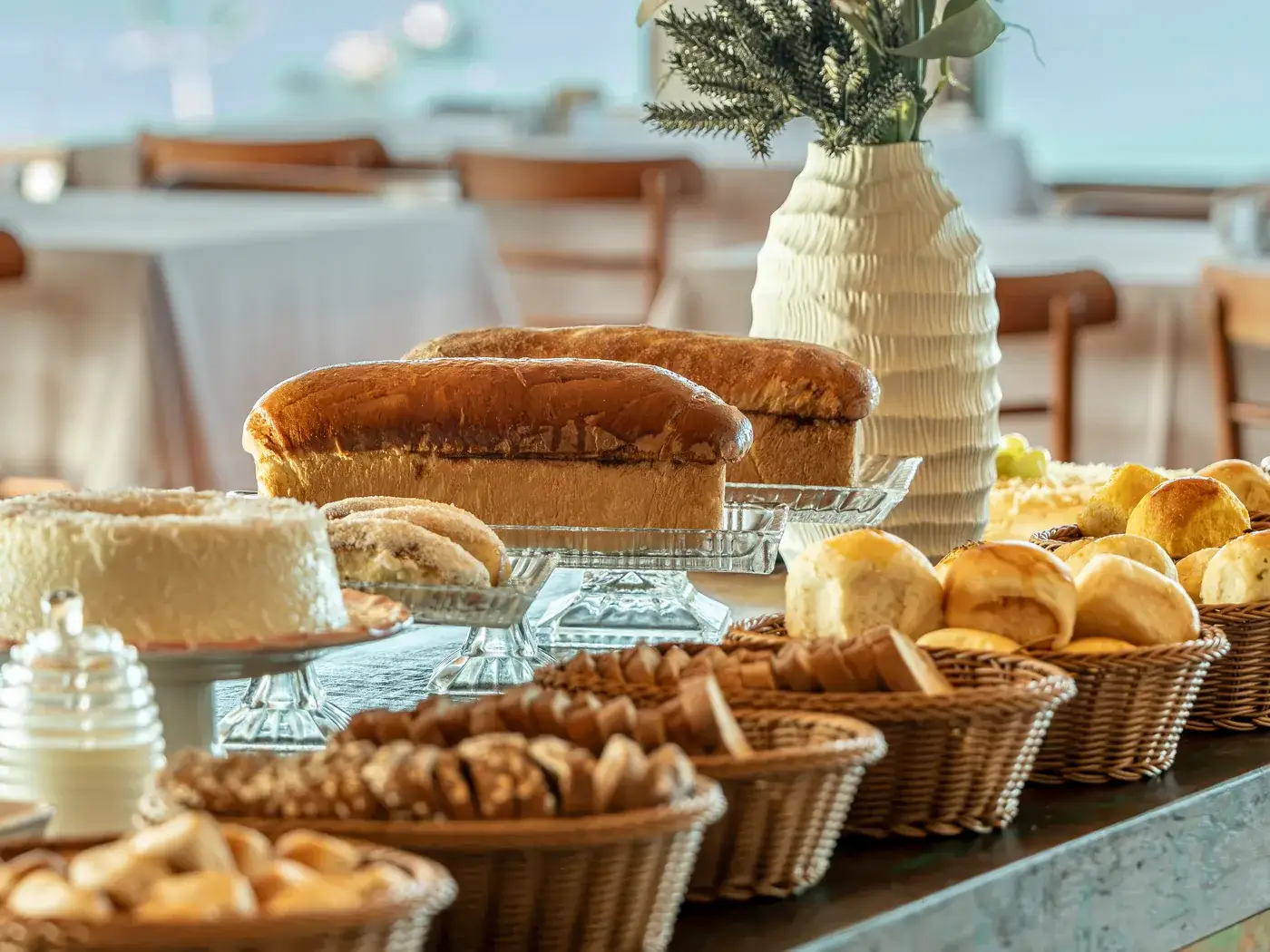 Pães caseiros, bolo e cestas de pão no buffet, com um vaso de flores e o mar ao fundo