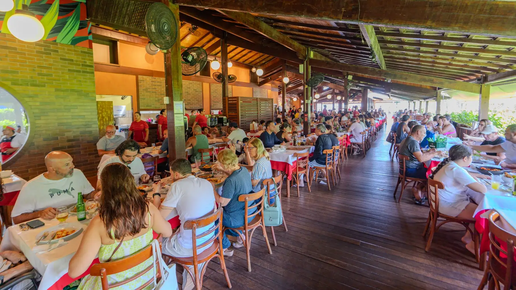 Salão do restaurante com cobertura de madeira, mesas ocupadas e o mar ao fundo