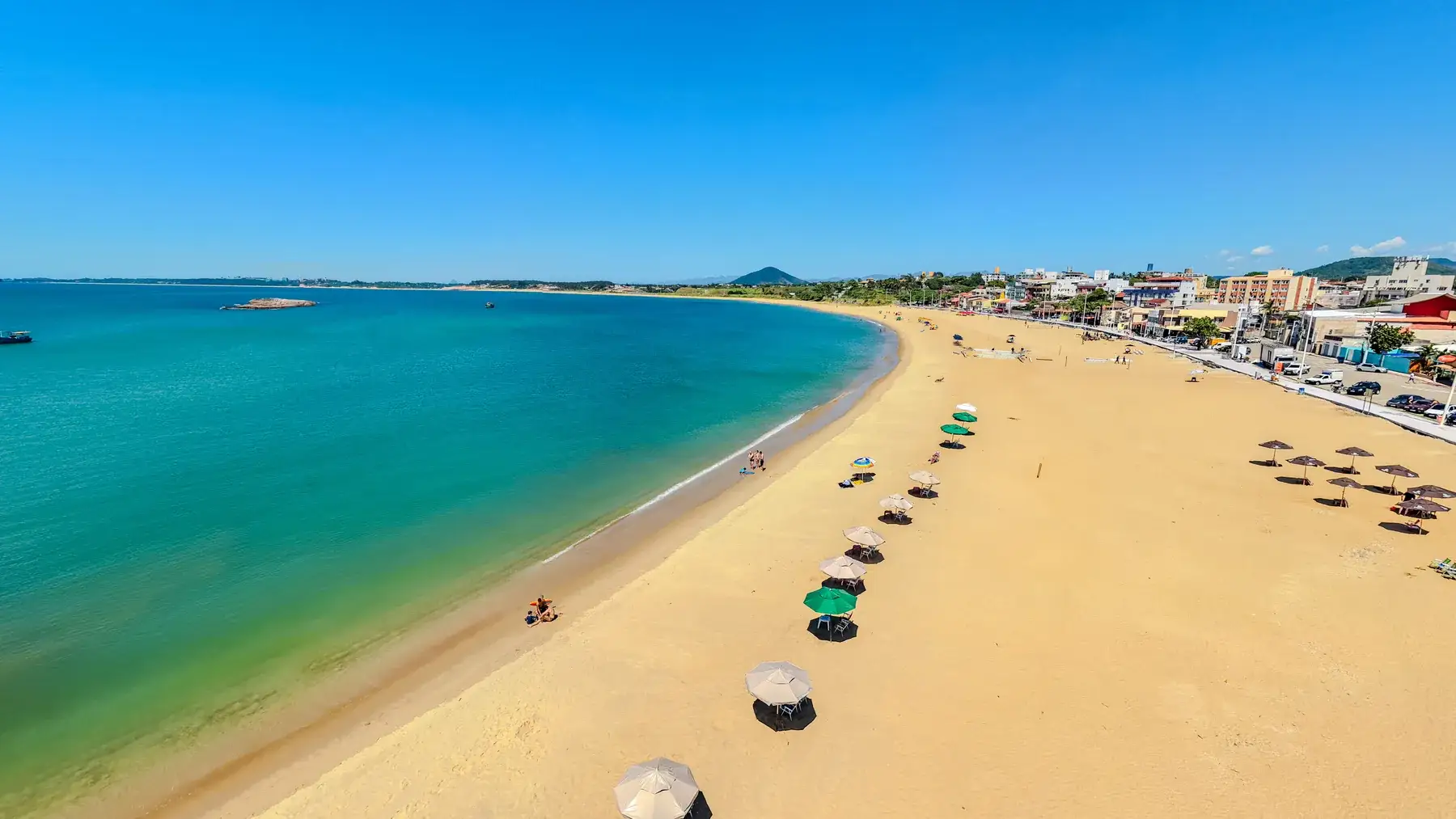 Vista aérea da curva inteira da Praia de Meaípe, com os guarda-sóis na areia, o mar turquesa e a vila baixa à direita