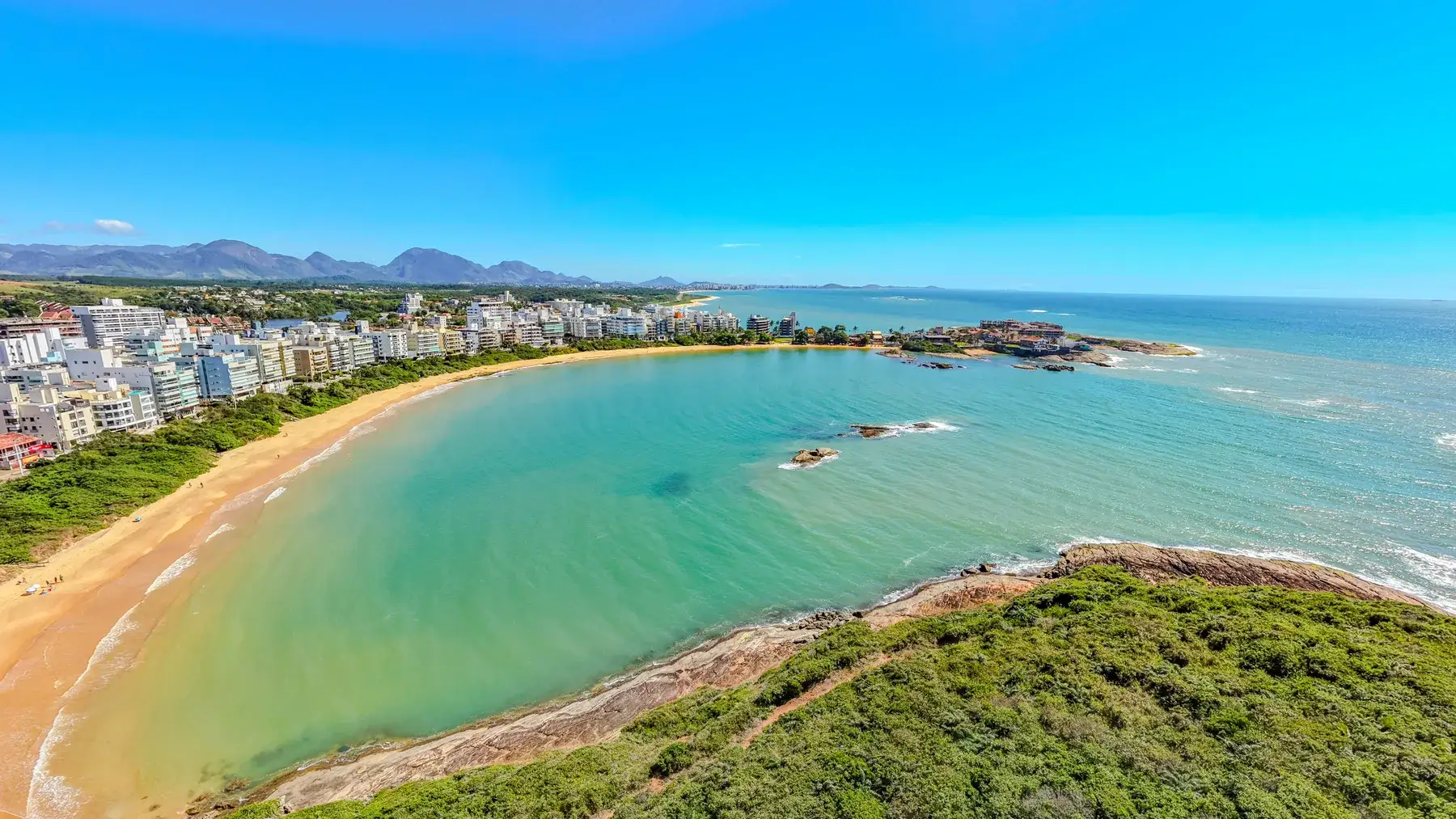 A curva longa da Praia de Peracanga vista do alto do costão, com o mar calmo, os prédios na orla e as montanhas ao fundo