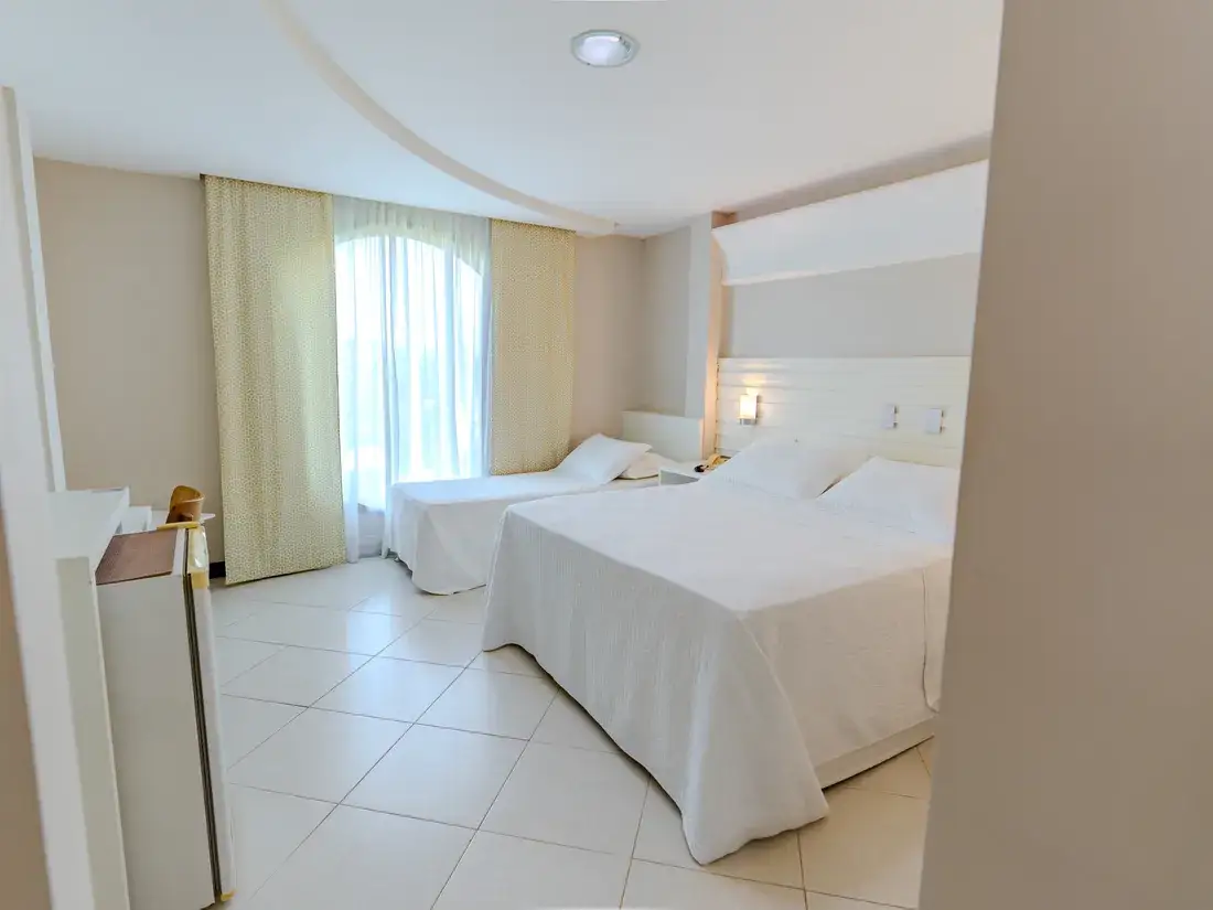 Quarto triplo visto da entrada, com o frigobar em primeiro plano, a cama de solteiro junto à janela e a queen ao lado