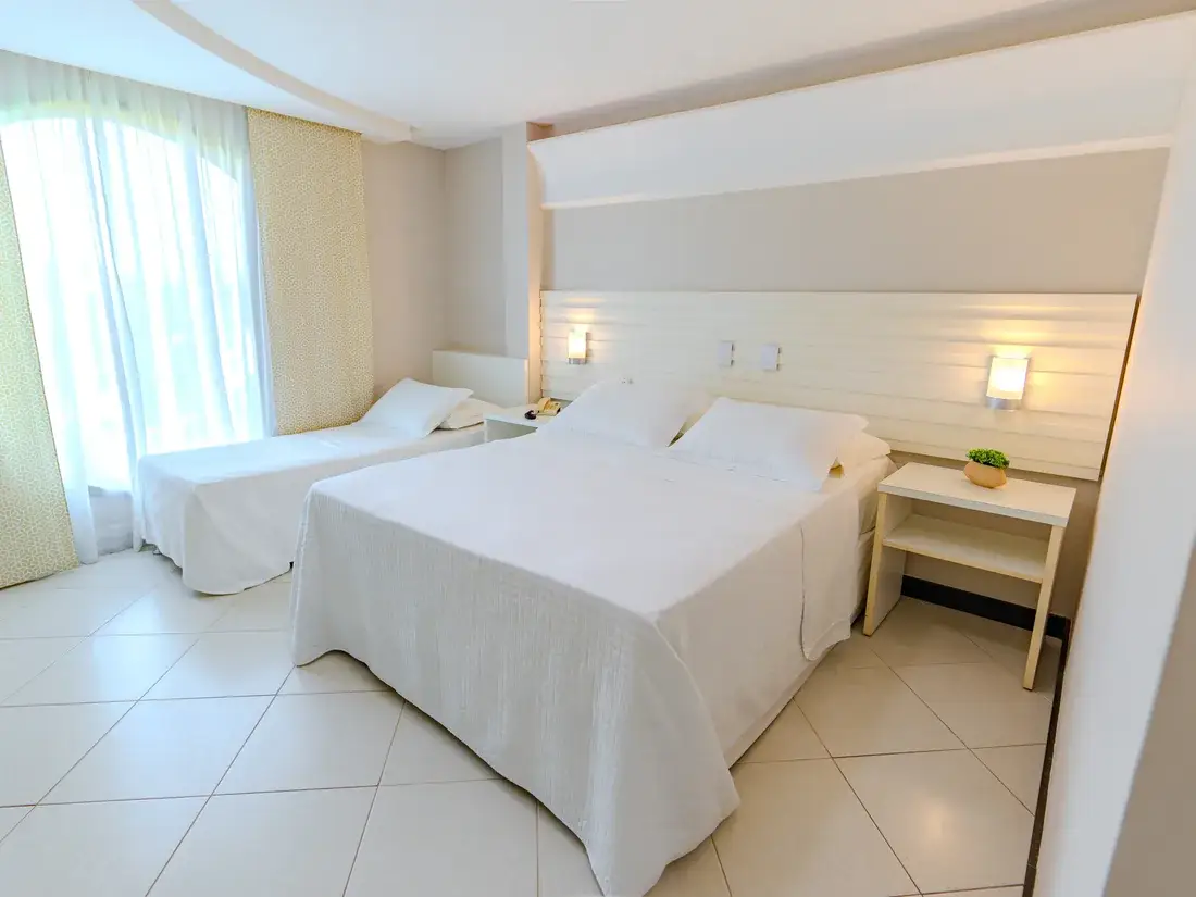 Quarto triplo com cama queen e cama de solteiro, luminárias acesas e luz entrando pela cortina